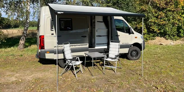 Fahrzeug von der Seite mit ausgefahrener Markise, darunter zwei Campingstühle und ein Tisch, im Hintergrund Birken und ein Feld